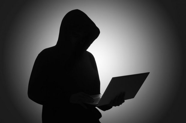 Hooded figure working on a laptop in a darkened room.